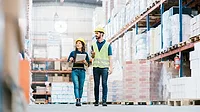 Two employees in hardhats checking inventory on warehouse racks.