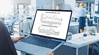 An engineer holds a laptop with mock-up charts. In the blurred background, three people are working together at a desk.