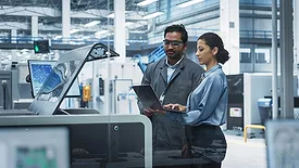 Male Industrial Engineer And Female Manager Using Laptop And Talking At Electronics Factory. The man is using a soldering jet printer.