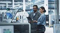 Male Industrial Engineer And Female Manager Using Laptop And Talking At Electronics Factory. The man is using a soldering jet printer.