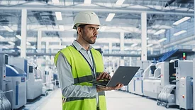 A male engineer is standing in a factory hall, using a laptop at a machinery manufacturing facility.