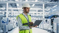 A male engineer is standing in a factory hall, using a laptop at a machinery manufacturing facility A male engineer is standing in a factory hall, using a laptop at a machinery manufacturing facility.