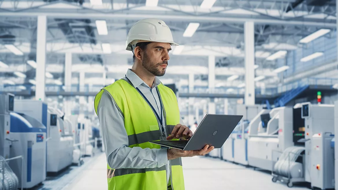 A male engineer is standing in a factory hall, using a laptop at a machinery manufacturing facility.