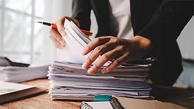 Person searching a stack of papers while holding a pen.