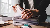 Person searching a stack of papers while holding a pen.