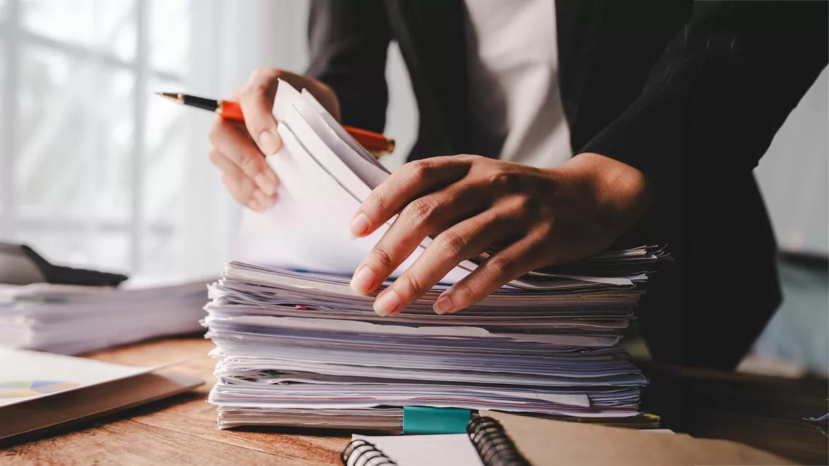 Person searching a stack of papers while holding a pen.