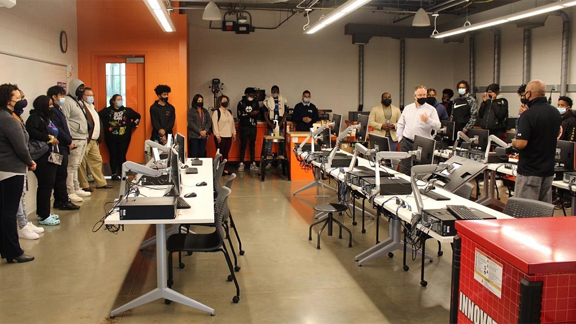 Students standing by the wall in a classroom listening to instructors.  The desks all have computers on them.  Everyone is wearing a mask.