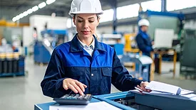 Female engineer wearing a white safety helmet in a factory.  A worker is behind her in the blurred background.