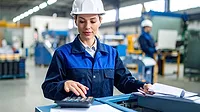 Female engineer wearing a white safety helmet in a factory.  A worker is behind her in the blurred background.