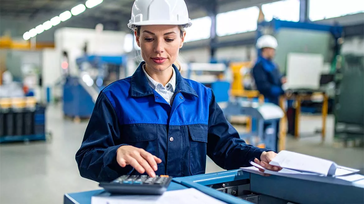 Female engineer wearing a white safety helmet in a factory. A worker is behind her in the blurred background.
