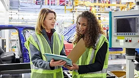 Car engineers in yellow safety vests discuss a file in a factory.