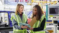 Car engineers discussing a file in a factory Car engineers in yellow safety vests discuss a file in a factory.