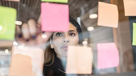Young woman is posting a pink post-it note on a glass board where several multicolored post-its are already up.