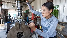 Female mechanic operating a milling machine in a garage workshop, wearing safety goggles, showcasing women in skilled trades and engineering.
