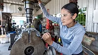 Female mechanic operating a milling machine in a garage workshop, wearing safety goggles, showcasing women in skilled trades and engineering.