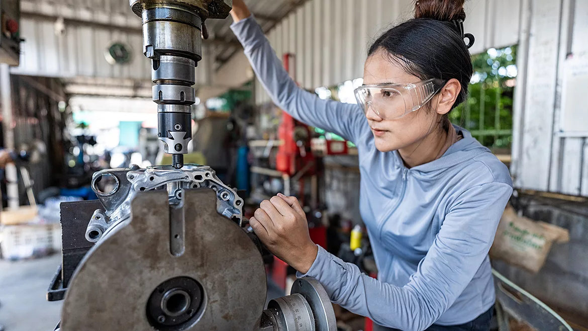 Female mechanic operating a milling machine in a garage workshop, wearing safety goggles, showcasing women in skilled trades and engineering.