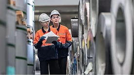 Two uniformed workers wearing safety hats and carrying out an inventory audit at a metal sheet factory.