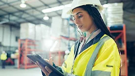 Female engineer in hard hat and safety jacket, working on a tablet in a warehouse.