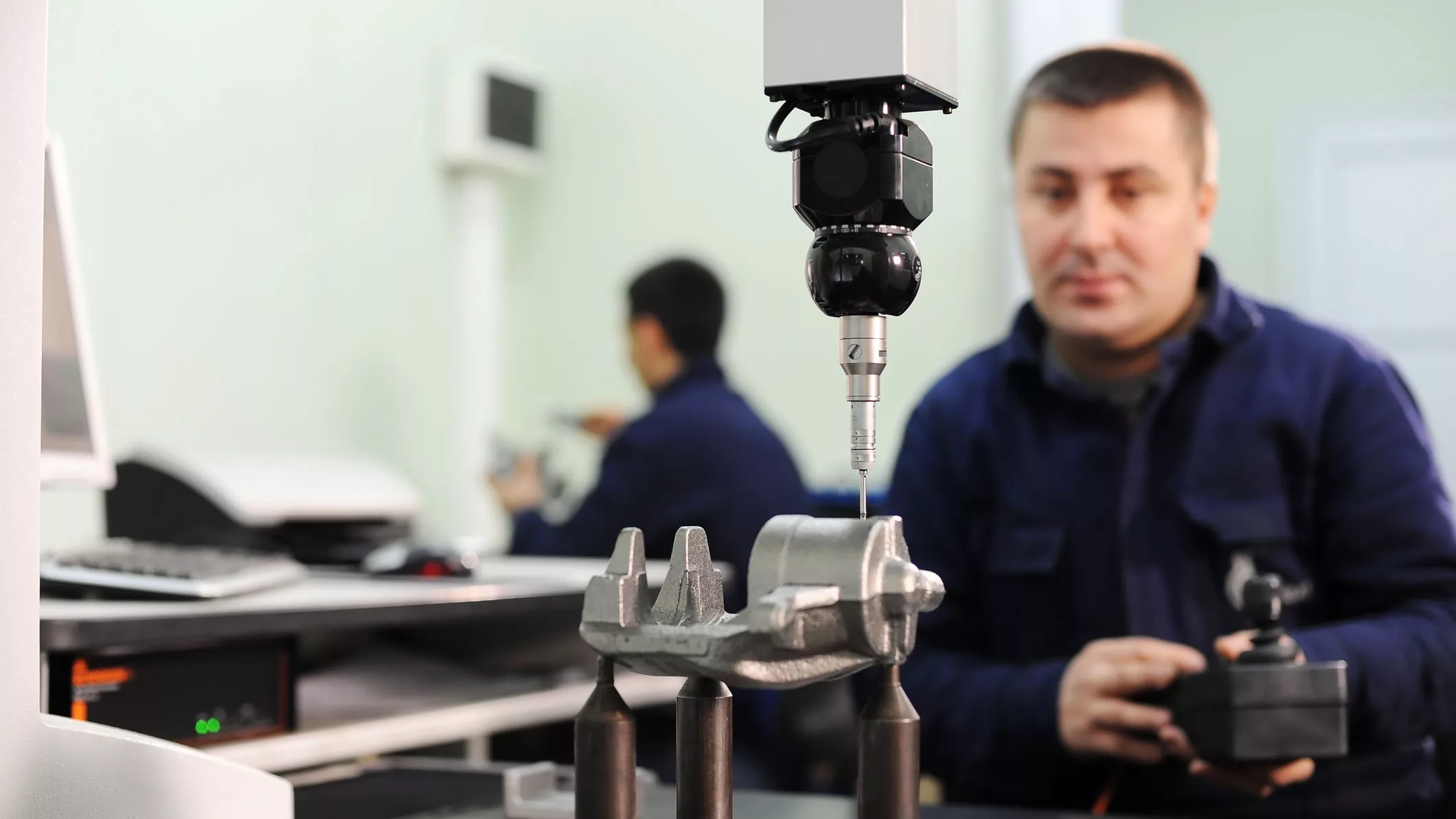 Worker is checking quality of new machine part. Another worker is in the blurred background.