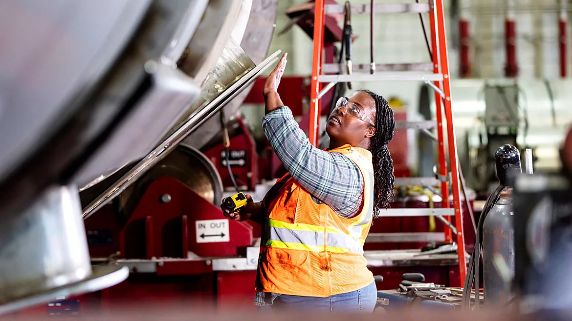 An African American woman working in a metal fabrication shop. She is wearing a safety vest and protective goggles, holding a tape measure and examining part of a large metal object.