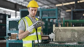 A male warehouse worker wearing a safety uniform and a helmet inspects the quality of products with a laptop computer in a storage warehouse.