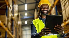Smiling warehouse worker in a reflective vest and hard hat checking inventory data on a tablet device in an organized storage facility.