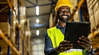 Smiling warehouse worker in a reflective vest and hard hat checking inventory data on a tablet device in an organized storage facility.
