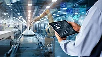 Back of a man holding a tablet monitoring a robotic arm in a factory.