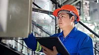 An industrial engineer wearing a hard hat and protective workwear examines machinery in a factory setting using a tablet.
