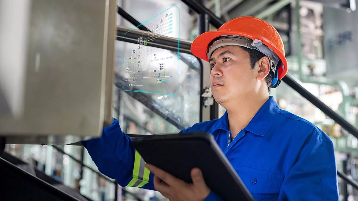 An industrial engineer wearing a hard hat and protective workwear examines machinery in a factory setting using a tablet.