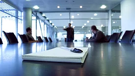Three men in a boardroom, one man pointing. Focus on notepad