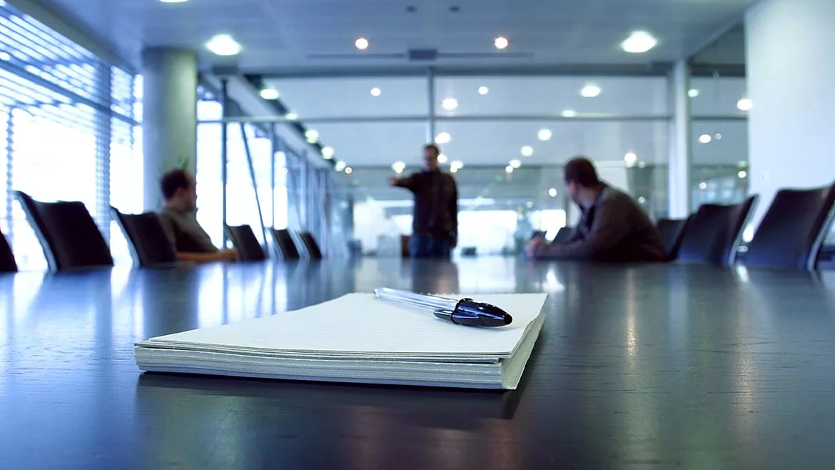 Three men in a boardroom, one man pointing. Focus on notepad