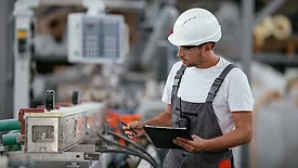 Male engineer in factory, wearing a white safety hardhat, and overalls.  The factory floor background is blurred.