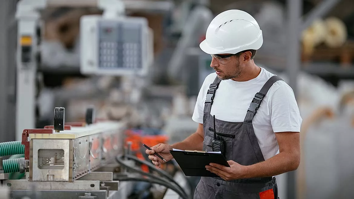 Male engineer in factory, wearing a white safety hardhat, and overalls. The factory floor background is blurred.