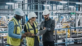 A team of automotive factory engineers in white safety hats and work vests are discussing something on a laptop and tablet. The assembly plant is slightly blurred in the background.