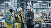 A team of automotive factory engineers in white safety hats and work vests are discussing something on a laptop and tablet. The assembly plant is slightly blurred in the background.