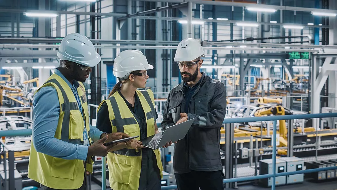 A team of automotive factory engineers in white safety hats and work vests are discussing something on a laptop and tablet. The assembly plant is slightly blurred in the background.