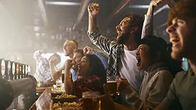a group of male friends in a sports bar, watching a game on a screen and celebrating. 