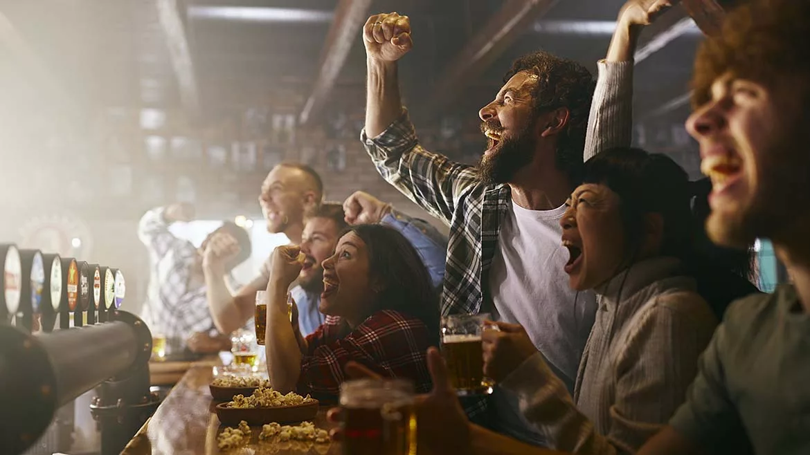 a group of male friends in a sports bar, watching a game on a screen and celebrating.