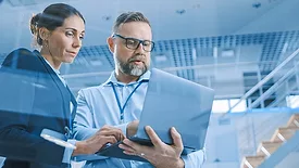Man and woman is standing while working on a laptop the man is holding. The background shows a set of modern stairs in an office building.