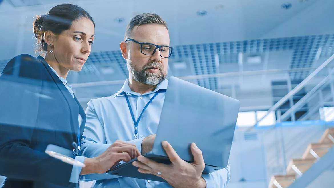 Man and woman is standing while working on a laptop the man is holding. The background shows a set of modern stairs in an office building.