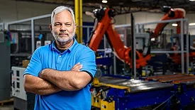 a man in a factory setting with a large orange KUKA industrial robot behind him