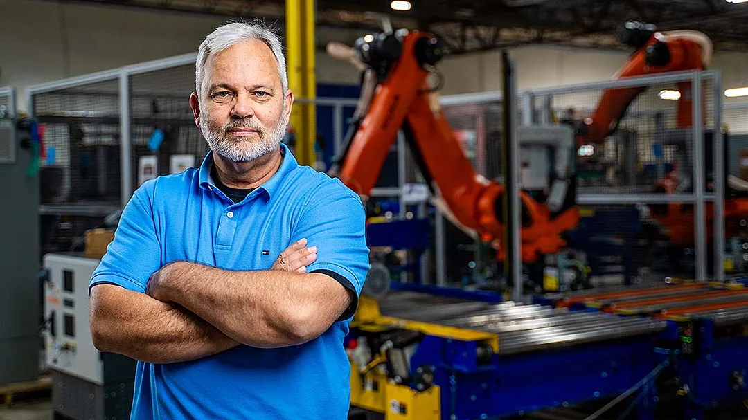 a man in a factory setting with a large orange KUKA industrial robot behind him