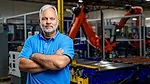 a man in a factory setting with a large orange KUKA industrial robot behind him