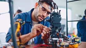 The image shows a man, likely an electronics technician or engineer, working on a complex electronic device or robot in a workshop environment