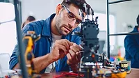 The image shows a man, likely an electronics technician or engineer, working on a complex electronic device or robot in a workshop environment
