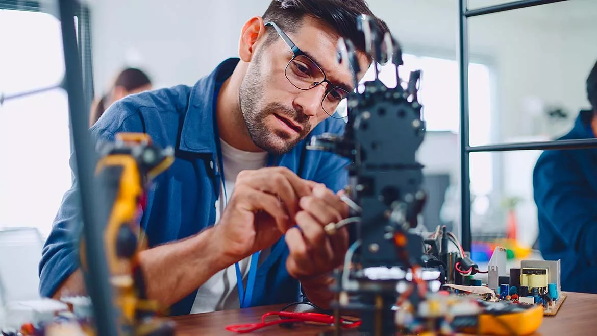 The image shows a man, likely an electronics technician or engineer, working on a complex electronic device or robot in a workshop environment