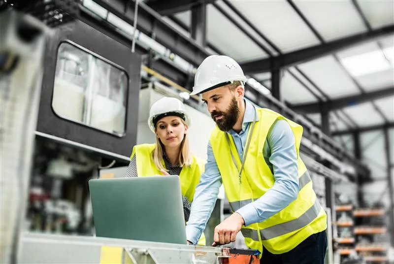 two industrial workers in a factory setting, using a laptop