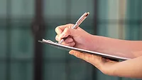 The image depicts a person writing on a clipboard The image depicts a person writing on a clipboard