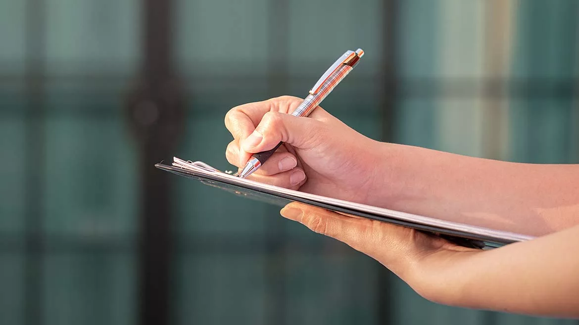 The image depicts a person writing on a clipboard